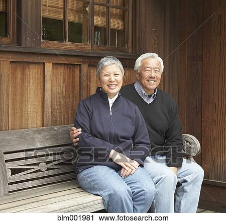 Stock Image - Senior Asian couple smiling and sitting on bench outdoors. Fotosearch