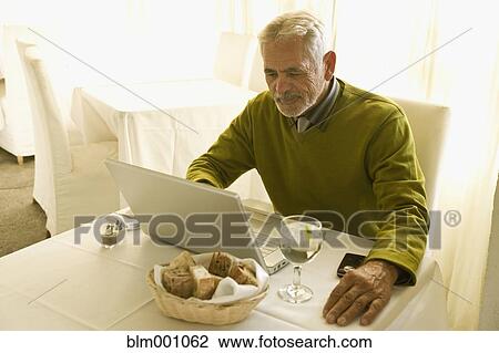 Stock Image - Senior Hispanic man using laptop at restaurant table. Fotosearch