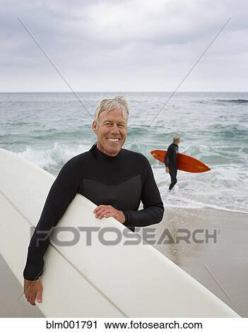 Stock Image - Senior man holding surfboard at beach. Fotosearch