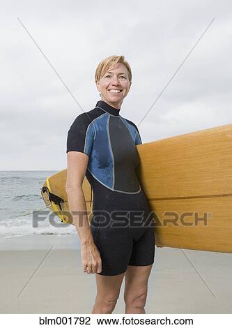 Stock Image - Senior woman holding surfboard at beach. Fotosearch