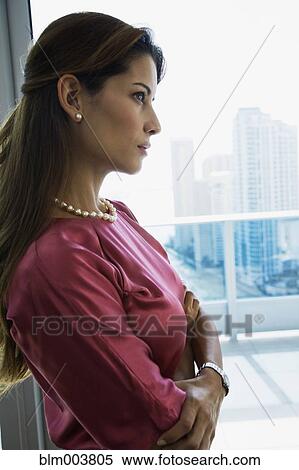 Stock Photography - Serious Hispanic woman standing by window with arms crossed. Fotosearch