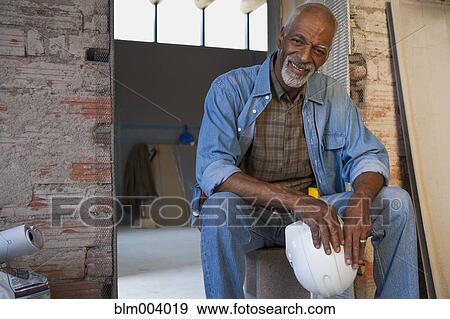 Stock Photo - Smiling African man holding hard-hat at construction site. Fotosearch