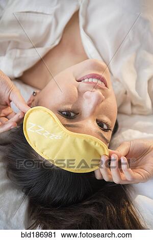 Stock Image - Smiling woman wearing eye mask on bed. Fotosearch