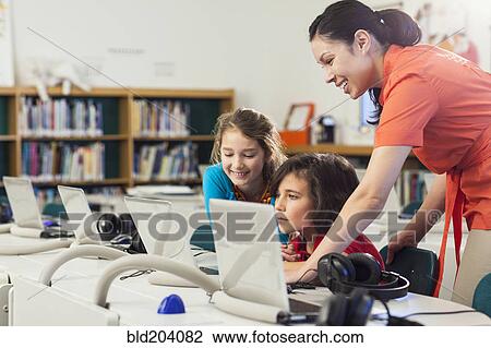 Stock Image - Teacher and children using laptop in library. Fotosearch