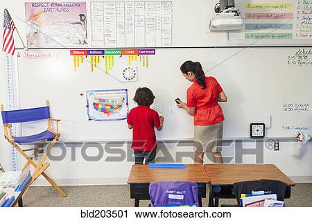 Stock Image - Teacher helping student at whiteboard in classroom. Fotosearch