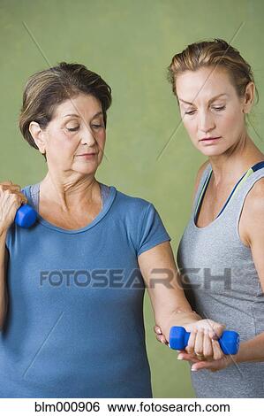 Stock Photograph - Trainer helping senior woman lift hand weights. Fotosearch