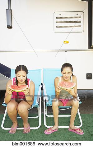 Stock Photograph - Two Hispanic sisters eating watermelon next to recreational vehicle. Fotosearch