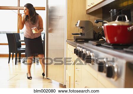 Stock Image - Woman eating ice cream next to refrigerator. Fotosearch