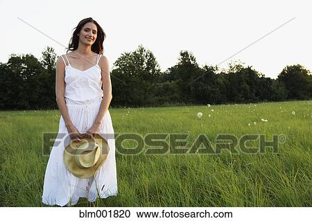 Stock Image - Woman standing in meadow holding straw hat. Fotosearch