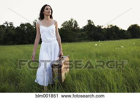 Stock Image - Woman walking with suitcase through meadow. Fotosearch