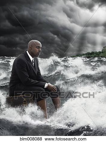 Stock Photo - African American businessman sitting on suitcase in stormy sea. Fotosearch