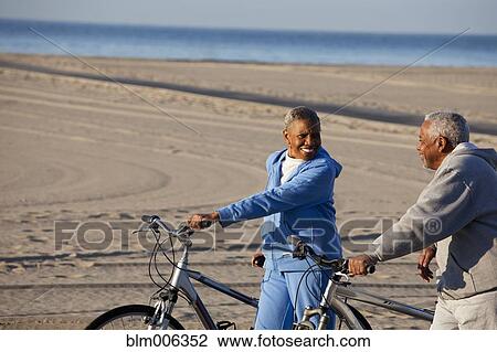 Stock Image - African American couple standing with bicycles on beach. Fotosearch