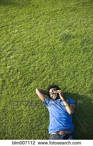 Stock Image - African American man using cell phone laying on grass. Fotosearch