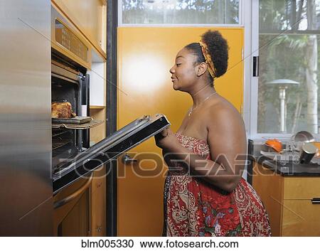 Stock Image - African American woman checking on food in oven. Fotosearch