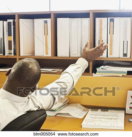 Picture - African businessman reaching for binder in office. Fotosearch