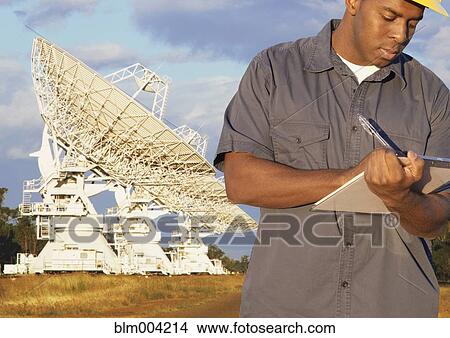 Picture - African man writing on clipboard with radio telescopes in background. Fotosearch