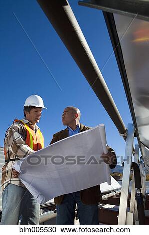 Stock Image - Architect and construction worker on rooftop near solar panels. Fotosearch