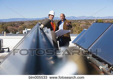 Stock Image - Architect and construction worker on rooftop near solar panels. Fotosearch