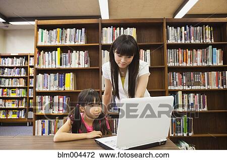 Stock Image - Asian woman helping girl use laptop in library. Fotosearch