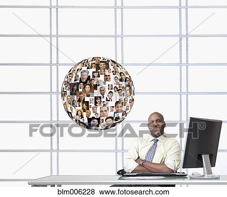 Stock Photo - Black businessman at desk with floating globe of faces. Fotosearch