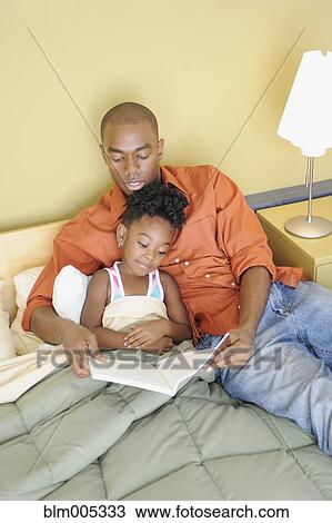 Stock Image - Black father reading book to daughter in bedroom. Fotosearch