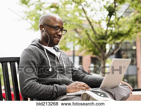 Black man using laptop and earphones on city bench View Large Photo Image Stock Photo - Black man using laptop and earphones on city bench. Fotosearch