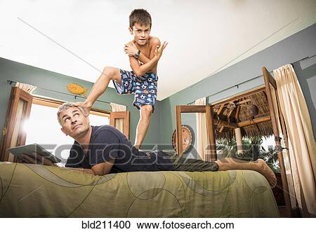 Stock Image - Boy standing on father on bed. Fotosearch