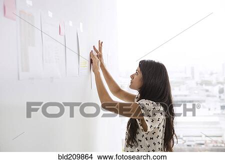 Picture - Businesswoman taping up papers on office wall. Fotosearch
