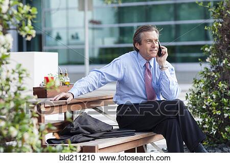 Stock Image - Caucasian businessman on cell phone on picnic bench. Fotosearch