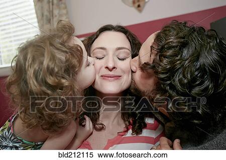 Stock Photography - Caucasian father and daughter kissing mother's cheeks in bedroom. Fotosearch