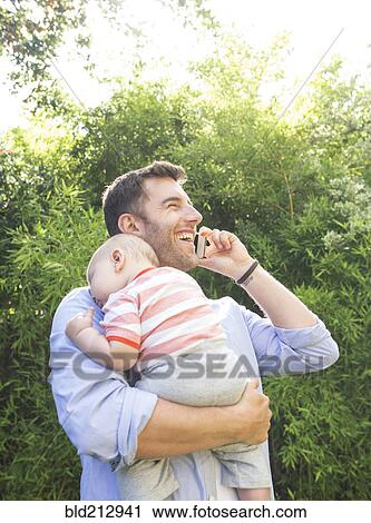 Stock Image - Caucasian father on cell phone holding baby. Fotosearch