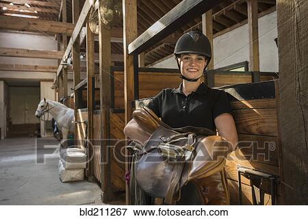 Stock Photo - Caucasian girl carrying saddle in horse stable. Fotosearch