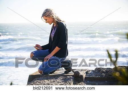 Stock Image - Caucasian girl using cell phone on hilltop overlooking ocean. Fotosearch