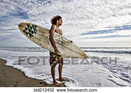 Stock Image - Caucasian man holding surfboard in waves on beach. Fotosearch