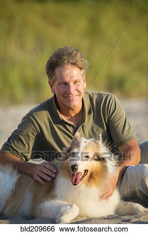 Stock Photograph - Caucasian man sitting with dog on beach. Fotosearch