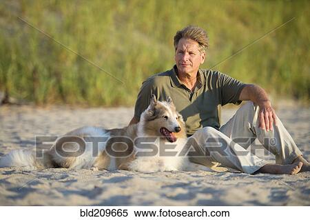 Stock Photography - Caucasian man sitting with dog on beach. Fotosearch