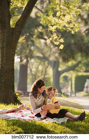 Stock Image - Caucasian mother reading story to daughter in park. Fotosearch