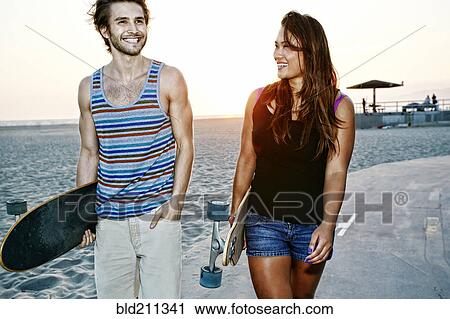 Stock Image - Couple carrying skateboards on beach. Fotosearch