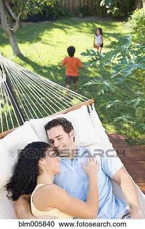 Stock Image - Couple relaxing in hammock with children playing in background. Fotosearch