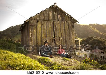 Stock Image - Couple sitting against barn in rural landscape. Fotosearch