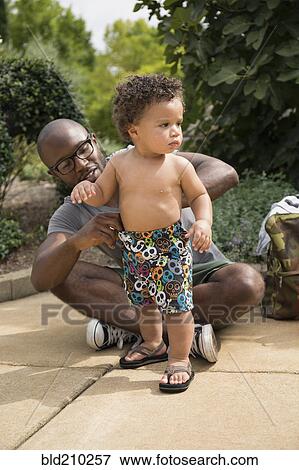 Stock Photo - Father adjusting toddler son's shorts in park. Fotosearch