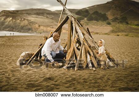 Stock Photo - Father and baby son sitting under driftwood teepee on beach. Fotosearch
