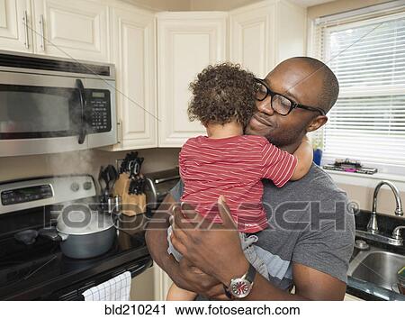 Stock Image - Father holding toddler son in kitchen. Fotosearch
