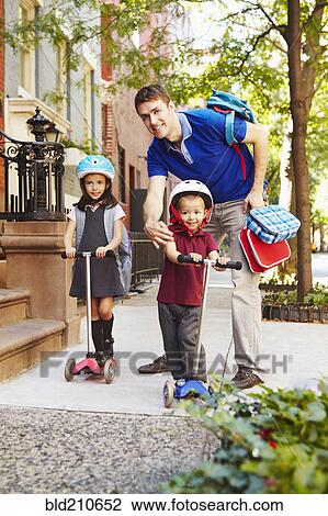 Stock Image - Father taking children on scooters to school. Fotosearch