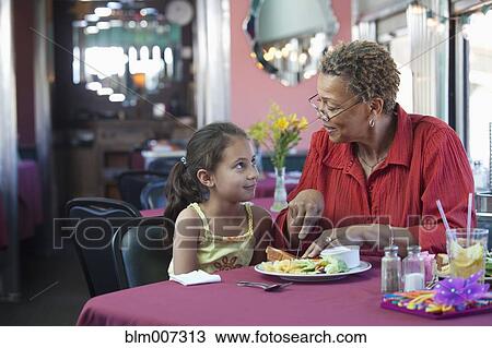 Stock Image - Grandmother cutting sandwich for granddaughter in restaurant. Fotosearch