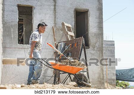 Stock Photo - Hispanic construction worker pushing wheelbarrow at construction site. Fotosearch