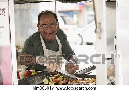 Stock Photo - Hispanic man grilling meat in food cart. Fotosearch