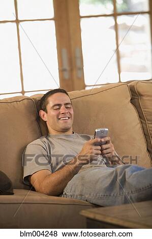 Stock Photo - Hispanic man text messaging with cell phone on sofa. Fotosearch