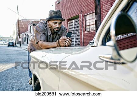 Stock Image - Hispanic man using cell phone on vintage car. Fotosearch