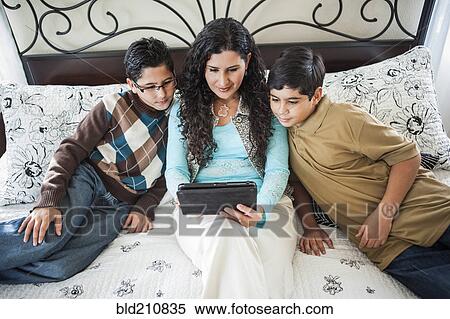 Stock Photography - Hispanic mother reading to sons on bed. Fotosearch